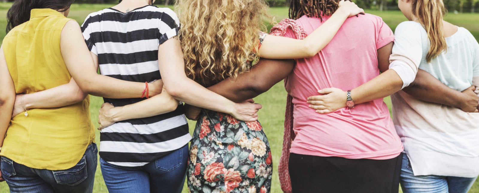 Back view of a diverse group of women locking arms and walking forward in the grass.