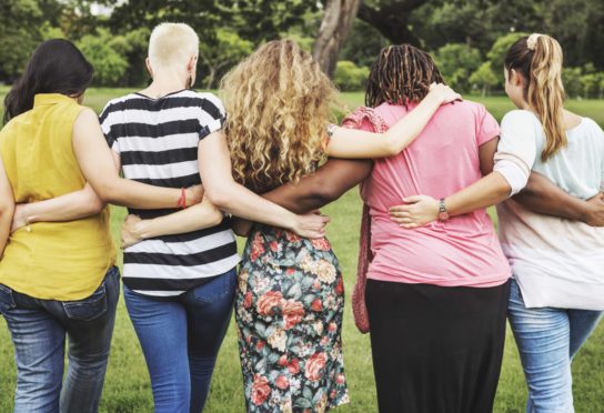 Back view of a diverse group of women locking arms and walking forward in the grass.