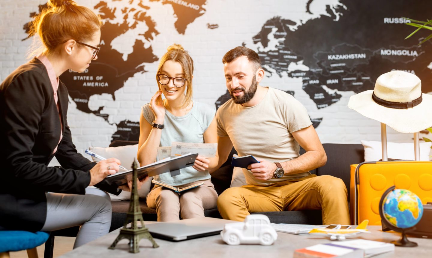 Couple sitting on a couch at a office talking to a lady with glasses holding a clipboard. In the background is a black and white world map wallpaper on the wall. To the right of the man on the couch is a yellow suitcase with a hat on top.