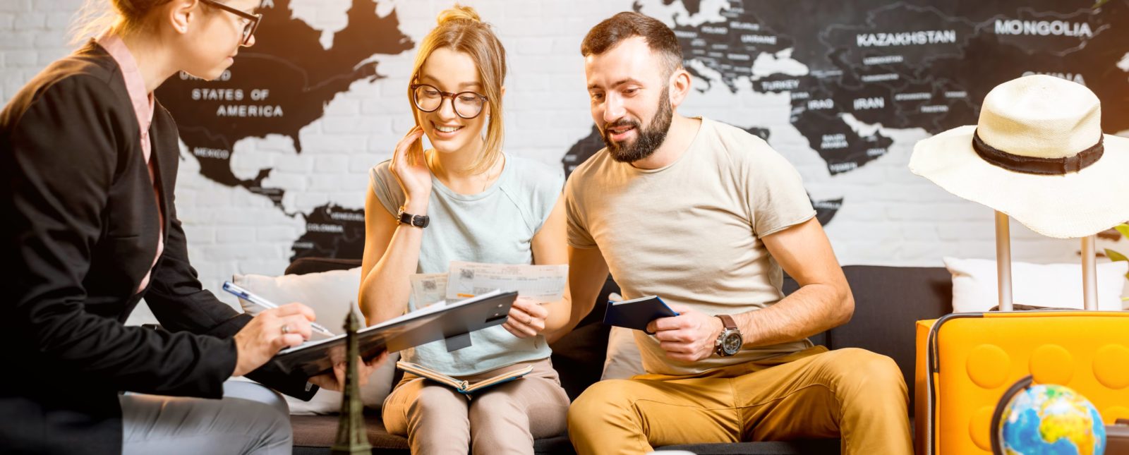 Couple sitting on a couch at a office talking to a lady with glasses holding a clipboard. In the background is a black and white world map wallpaper on the wall. To the right of the man on the couch is a yellow suitcase with a hat on top.