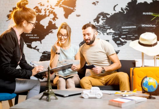 Couple sitting on a couch at a office talking to a lady with glasses holding a clipboard. In the background is a black and white world map wallpaper on the wall. To the right of the man on the couch is a yellow suitcase with a hat on top.