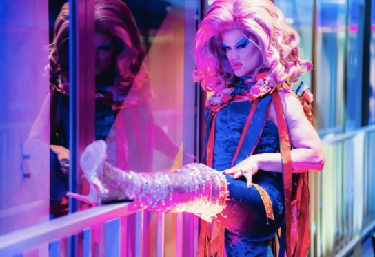 Drag queen wearing knee length boots, with leg up on a guard rail.