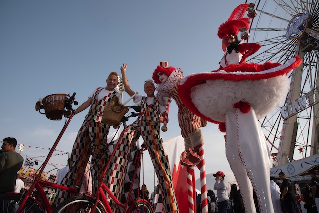 Clowns standing on stilts outside in a crowd of people with a carnival in the background