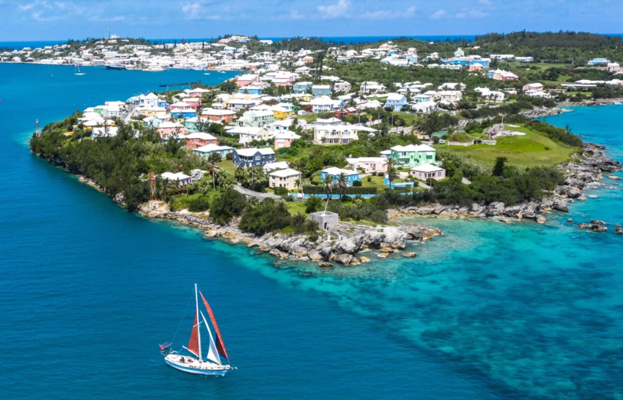 Buildings on island atop bright blue water, with blue and red sail boat in the foreground