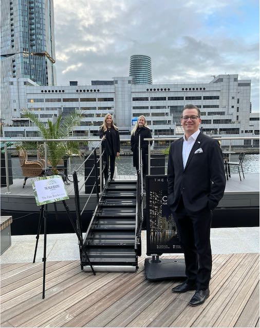 One man standing in front of Doc and two blonde women standing in the doc. All wearing all black.