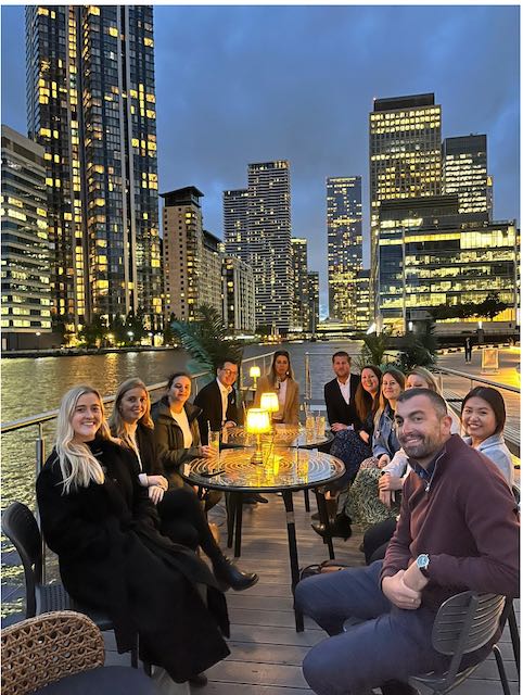 Team sitting in the evening having dinner on the docks, in front of city skyline.