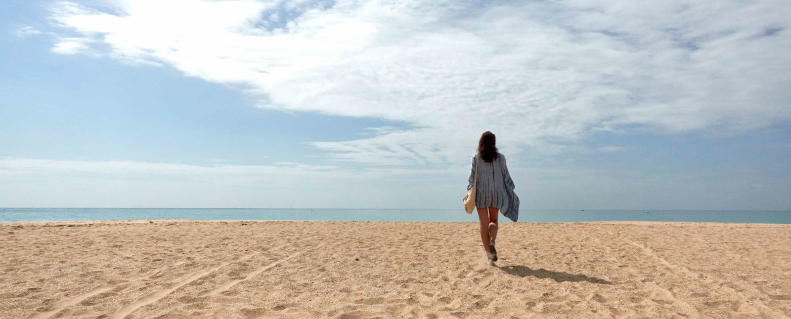 Woman walking away in the distance across a sandy tropical beach to the sea on a sunny day