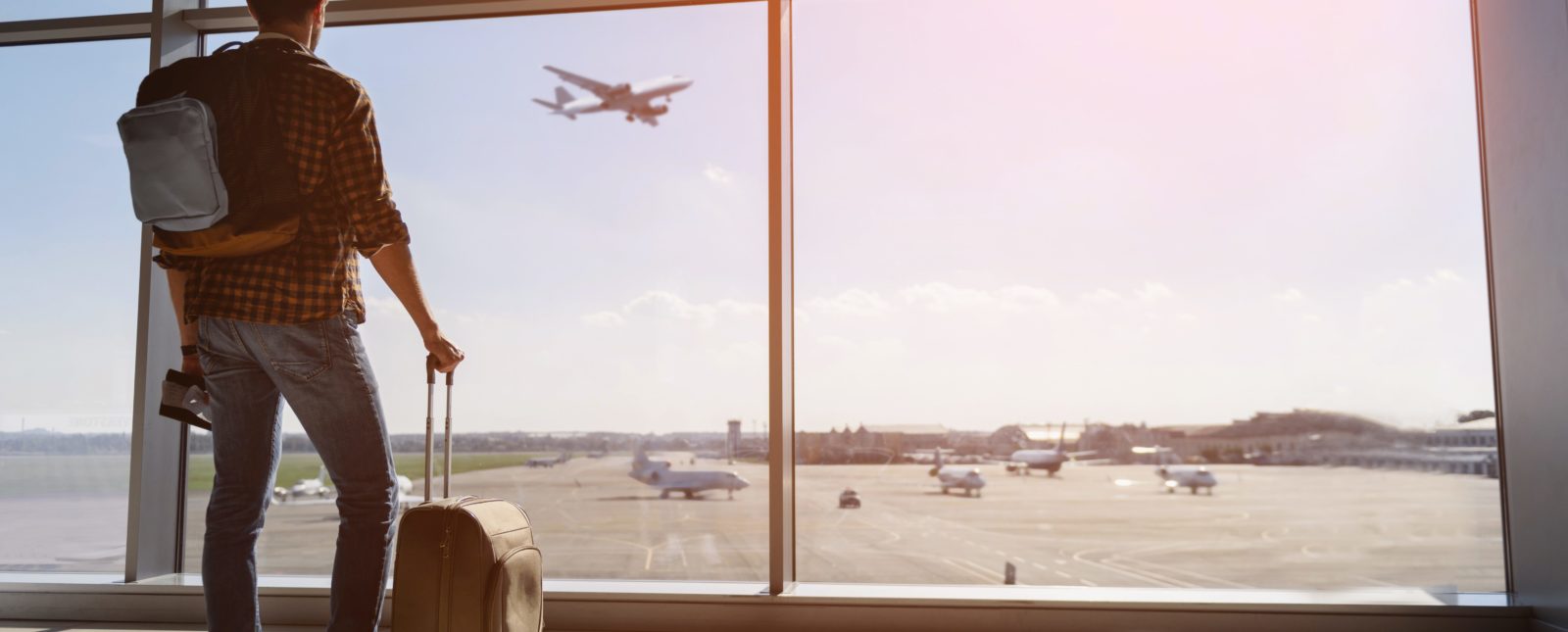 Calm male tourist is standing in airport and looking at aircraft flight through window. He is holding tickets and suitcase. Sunset