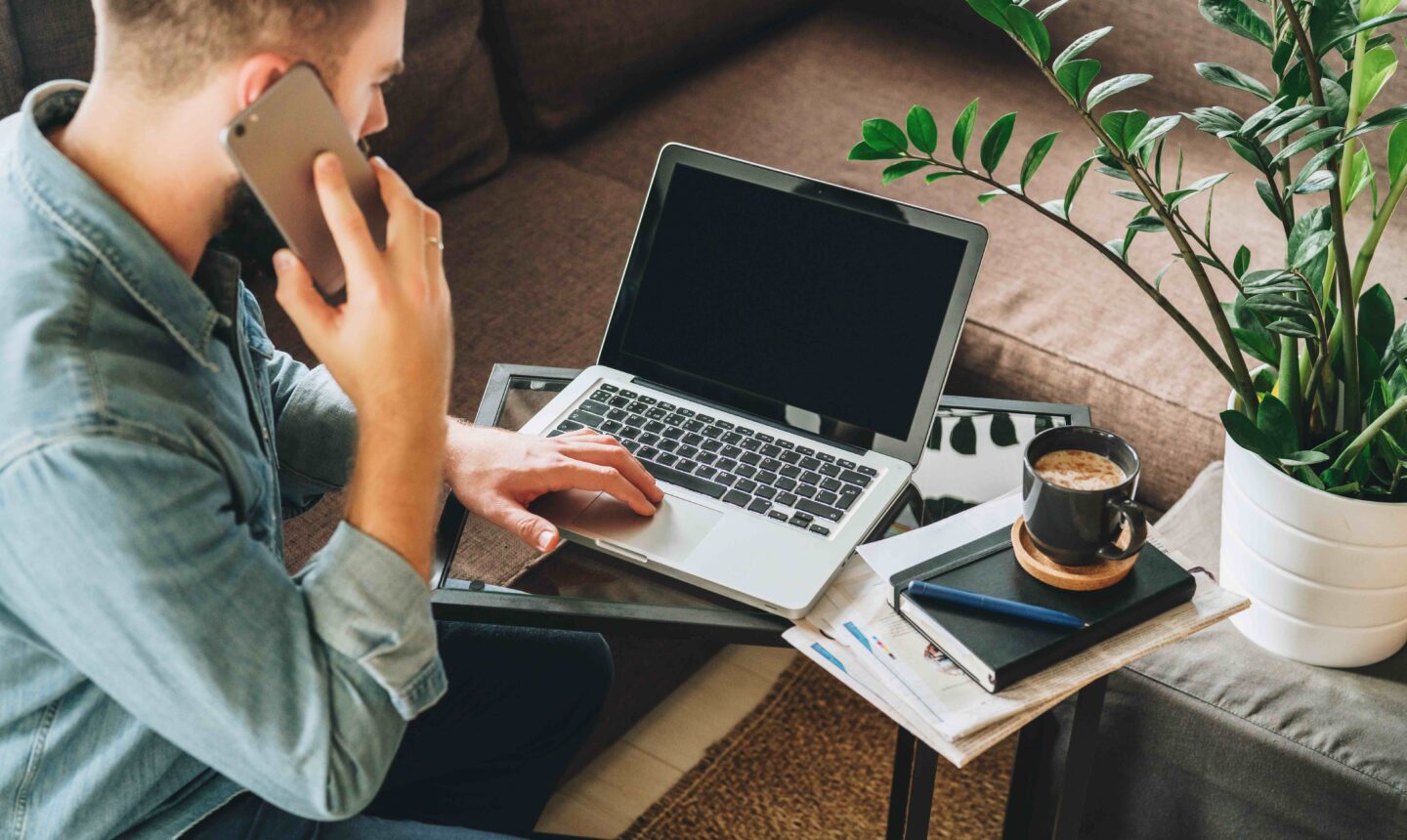 Back view. Young bearded hipster man, dressed in denim shirt, sits at home on couch at coffee table, uses laptop and talks on his cell phone, calling.