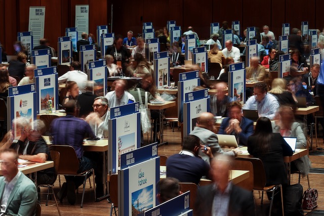 A blurred photo of a crowd of people sitting at individual tables for a conference.