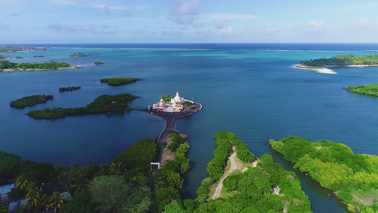 Overhead shot of multiple islands, greenery, a small building in the middle. Clear blue ocean.