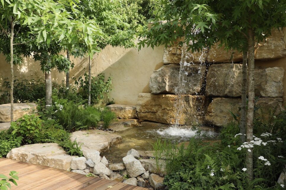 A small waterfall and pond with rocks behind it and a tree on a wooden platform.